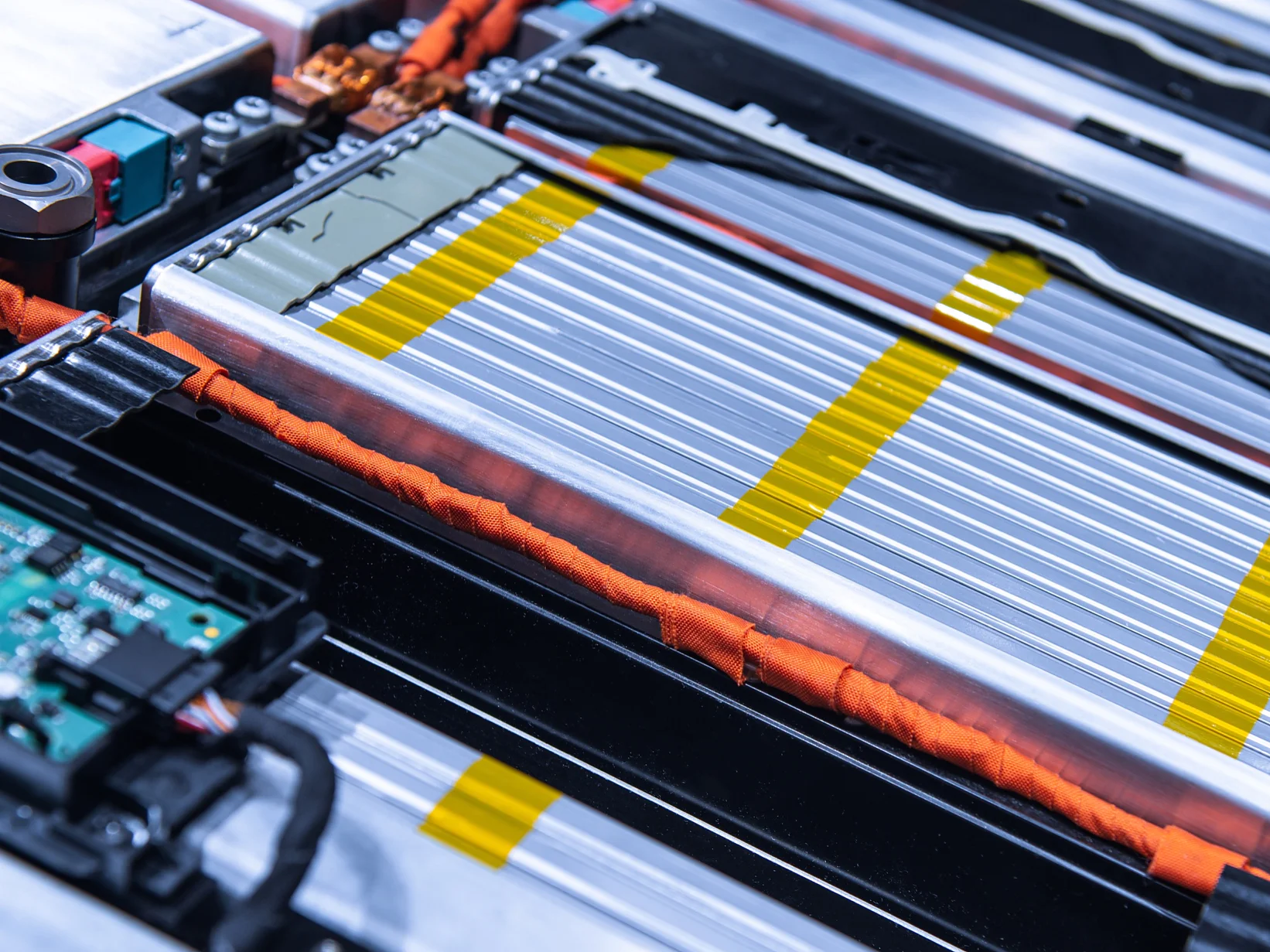 Close-up of a complex electrical circuit board and battery pack, featuring silver and yellow striped battery cells, orange wires, and a green circuit board. The components are tightly arranged within a metallic frame using tesa tape. (This text has been generated by AI)