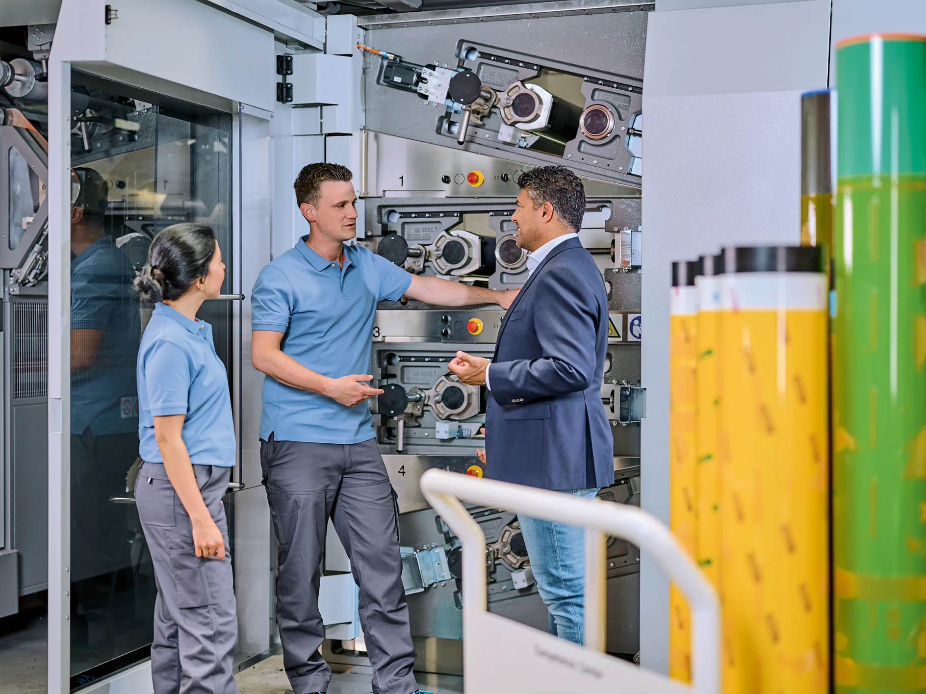 Flexo_Plate-mounting_Consultation_Bobst2023_002a-30646_V1 Three people stand in front of industrial machinery. Two individuals wear blue work uniforms, and one person in a blue blazer and jeans gestures while speaking. Rolls of tesa tape are visible in the foreground. (This text has been generated by AI)