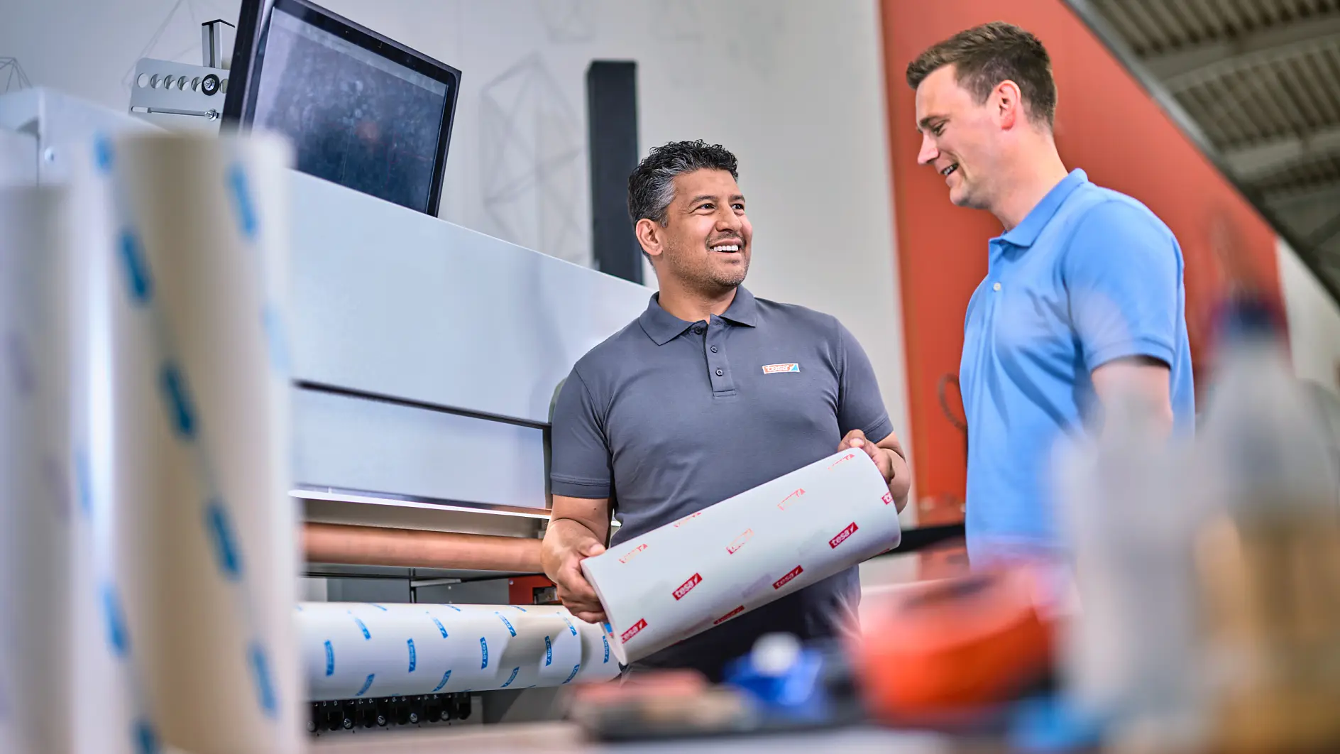 Two men are standing in a printing workshop. One man holds a roll of printed material secured with tesa tape, while the other looks on. A large printer and computer screen are in the background. Both men are dressed in casual work attire. (This text has been generated by AI)