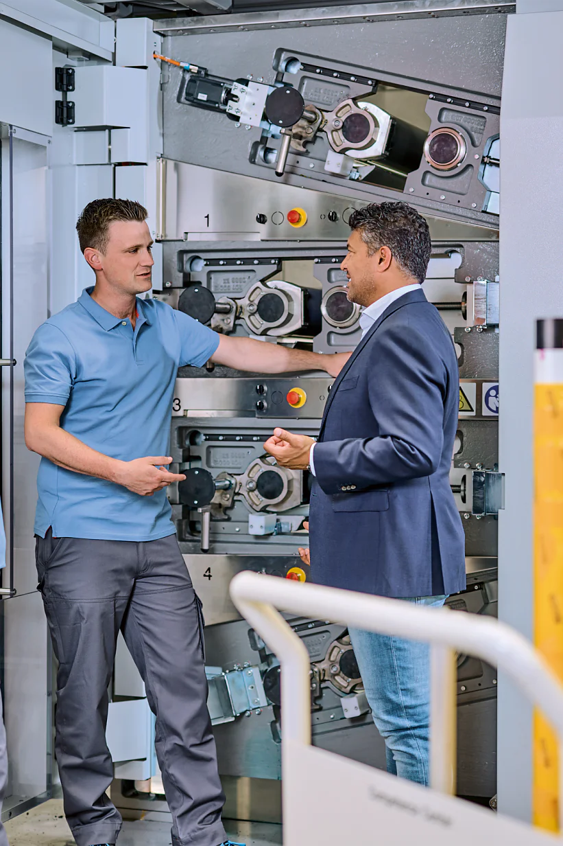 Three people stand in front of industrial machinery. Two individuals wear blue work uniforms, and one person in a blue blazer and jeans gestures while speaking. Rolls of tesa tape are visible in the foreground. (This text has been generated by AI)