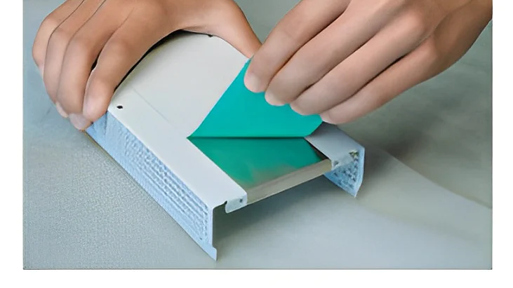 Duas mãos estão aplicando uma tira de fita tesa verde no interior de um invólucro plástico branco retangular. O invólucro apresenta um canal com uma superfície de metal, e as mãos estão posicionadas para prender a fita tesa suavemente. (This text has been generated by AI)