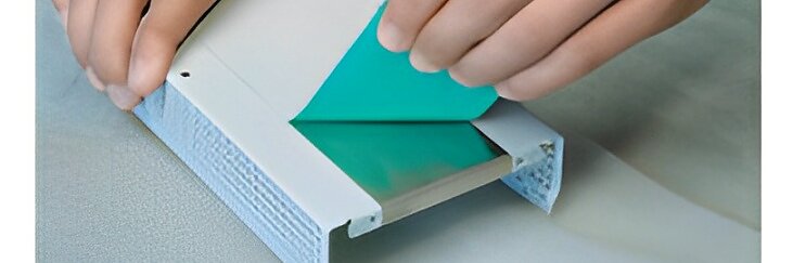 Duas mãos estão aplicando uma tira de fita tesa verde no interior de um invólucro plástico branco retangular. O invólucro apresenta um canal com uma superfície de metal, e as mãos estão posicionadas para prender a fita tesa suavemente. (This text has been generated by AI)