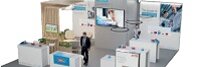 Um estande de exposição com vários cubículos brancos dispostos em formato de U. Um homem de terno anda pelo espaço. Uma tela grande exibe uma imagem acima da área central. Plantas em vasos decoram o estande, e a sinalização inclui logotipos e texto fixados com fita tesa. (This text has been generated by AI)