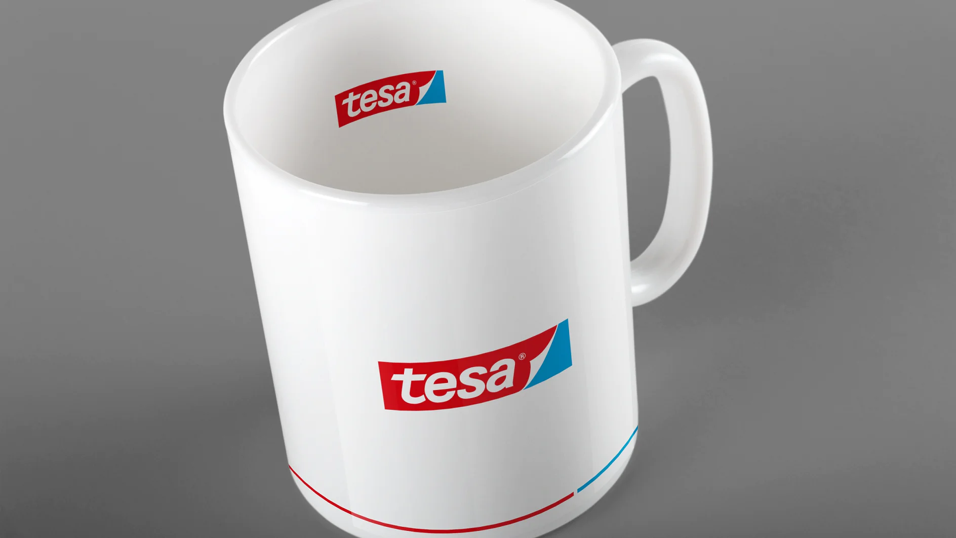 Uma caneca de cerâmica branca com alça é mostrada contra um fundo cinza. A caneca apresenta o logotipo tesa em vermelho e azul na lateral e na borda interna. Uma fina linha vermelha e azul circunda a base da caneca, exibindo um design elegante que incorpora os elementos de marca exclusivos da fita tesa. (This text has been generated by AI)