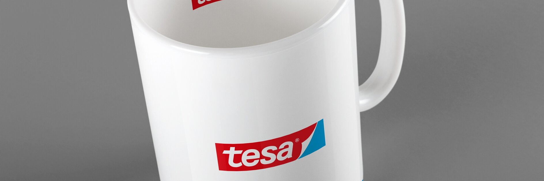 Uma caneca de cerâmica branca com alça é mostrada contra um fundo cinza. A caneca apresenta o logotipo tesa em vermelho e azul na lateral e na borda interna. Uma fina linha vermelha e azul circunda a base da caneca, exibindo um design elegante que incorpora os elementos de marca exclusivos da fita tesa. (This text has been generated by AI)