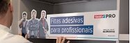 Uma pessoa está colocando uma placa em uma prateleira que diz Fitas adesivas para profissionais. A placa apresenta imagens de três pessoas e tem um logotipo no canto superior direito. Rolos de fita tesa são visíveis nas prateleiras acima. (This text has been generated by AI)