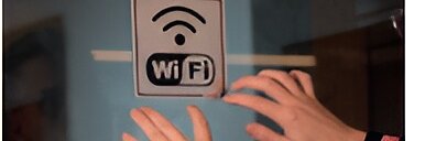 Duas mãos estão colocando ou ajustando uma placa em uma superfície de vidro usando fita tesa. A placa tem um símbolo de Wi-Fi e o texto WiFi exibido. (This text has been generated by AI)