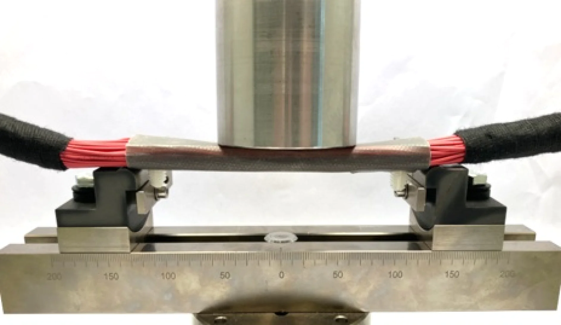 Um cilindro metálico comprime um feixe de fios vermelhos envoltos em fita tesa protetora. A configuração é mantida por dois grampos pretos em uma base de aço com uma escala de medição, indicando um processo de teste ou medição. (This text has been generated by AI)