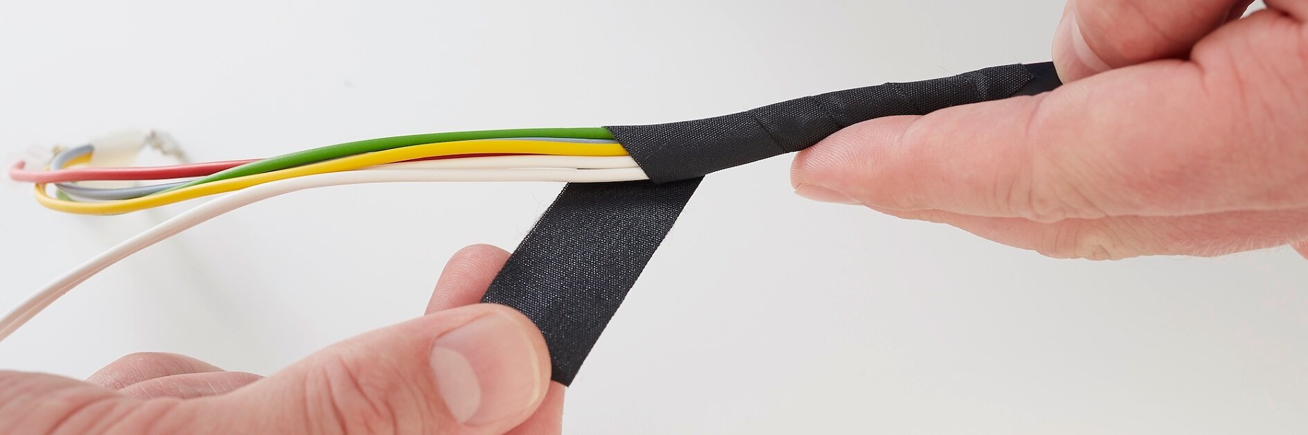 Close-up de mãos enrolando fita tesa preta em volta de um feixe de fios multicoloridos, incluindo branco, amarelo, verde e laranja. O fundo é uma superfície simples e clara. (This text has been generated by AI)