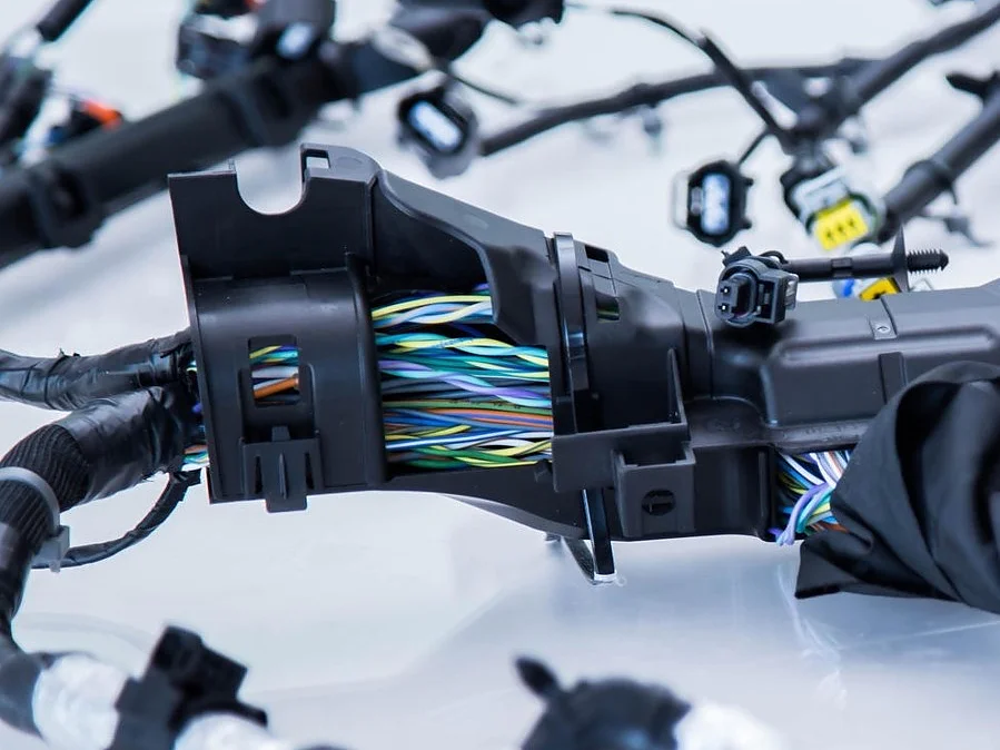 Close-up de um chicote de fiação complexo com vários fios multicoloridos agrupados e conectados com conectores de plástico preto, presos com fita tesa. A configuração repousa sobre uma superfície lisa e de cor clara. (This text has been generated by AI)