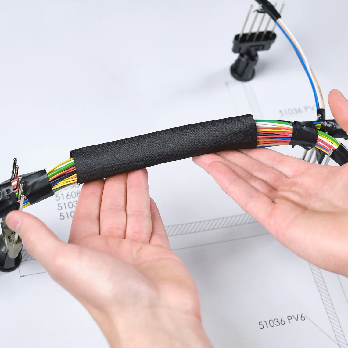 Duas mãos seguram um feixe de fios multicoloridos enrolados com uma capa protetora preta. Os fios são parte de um chicote elétrico automotivo, posicionado sobre um desenho técnico em uma mesa. Clipes de plástico prendem seções do chicote no fundo, enquanto a fita tesa garante que o feixe fique bem organizado. (This text has been generated by AI)