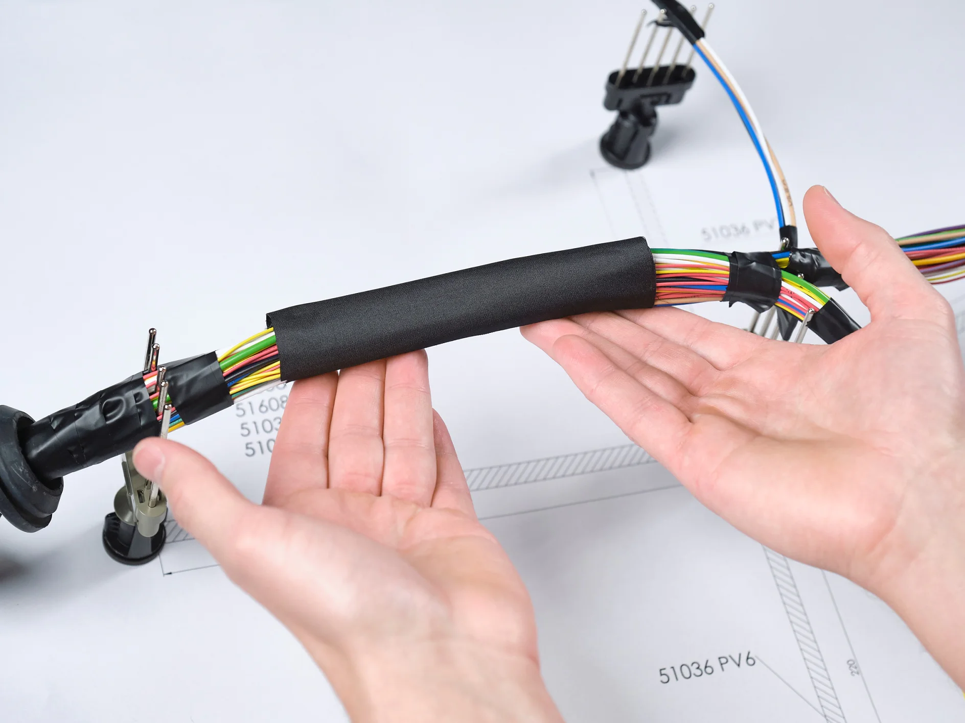Duas mãos seguram um feixe de fios multicoloridos enrolados com uma capa protetora preta. Os fios são parte de um chicote elétrico automotivo, posicionado sobre um desenho técnico em uma mesa. Clipes de plástico prendem seções do chicote no fundo, enquanto a fita tesa garante que o feixe fique bem organizado. (This text has been generated by AI)