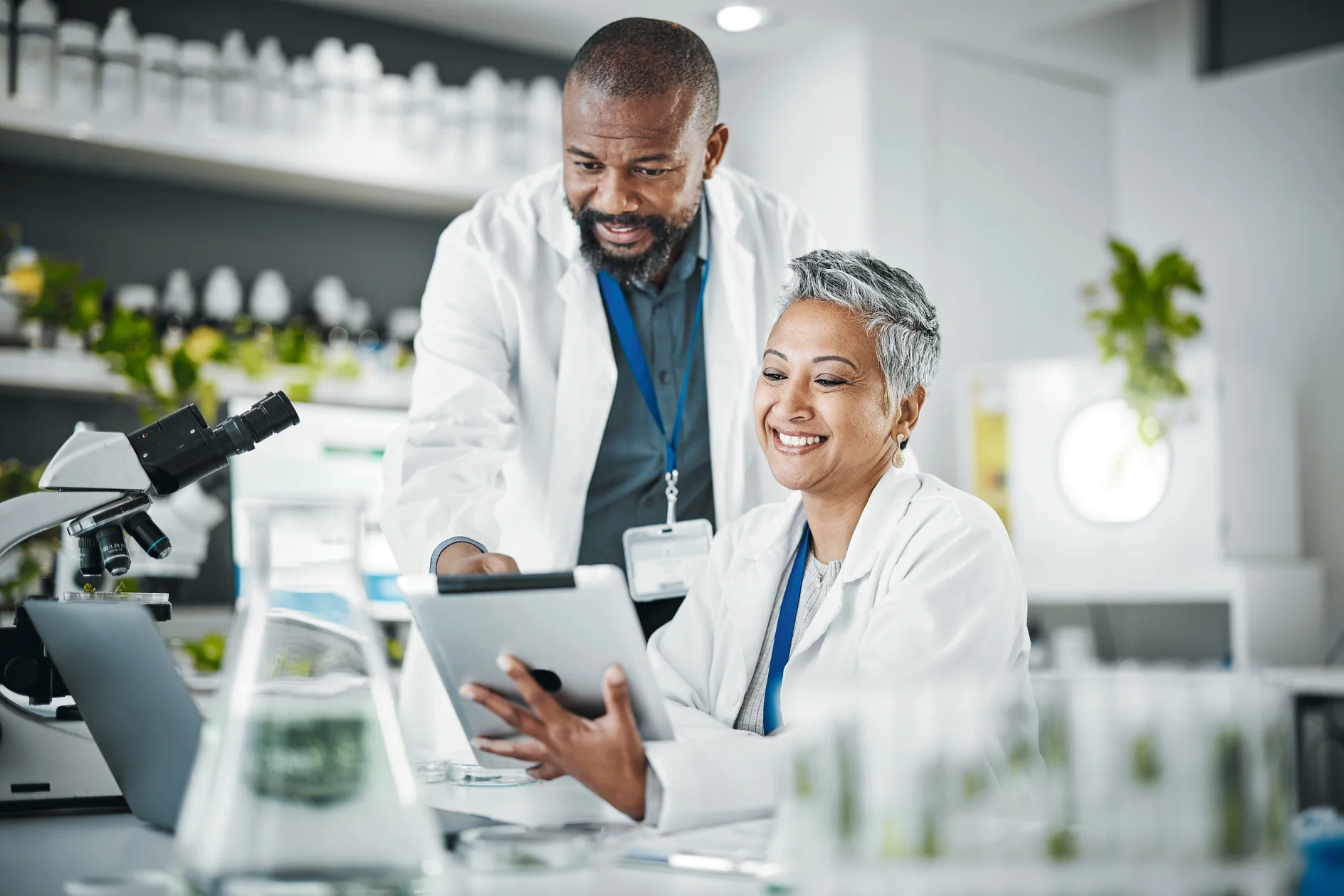Two scientists in white lab coats using a tablet in a bright laboratory with microscopes and glassware on the table tesa (This text has been generated by AI)