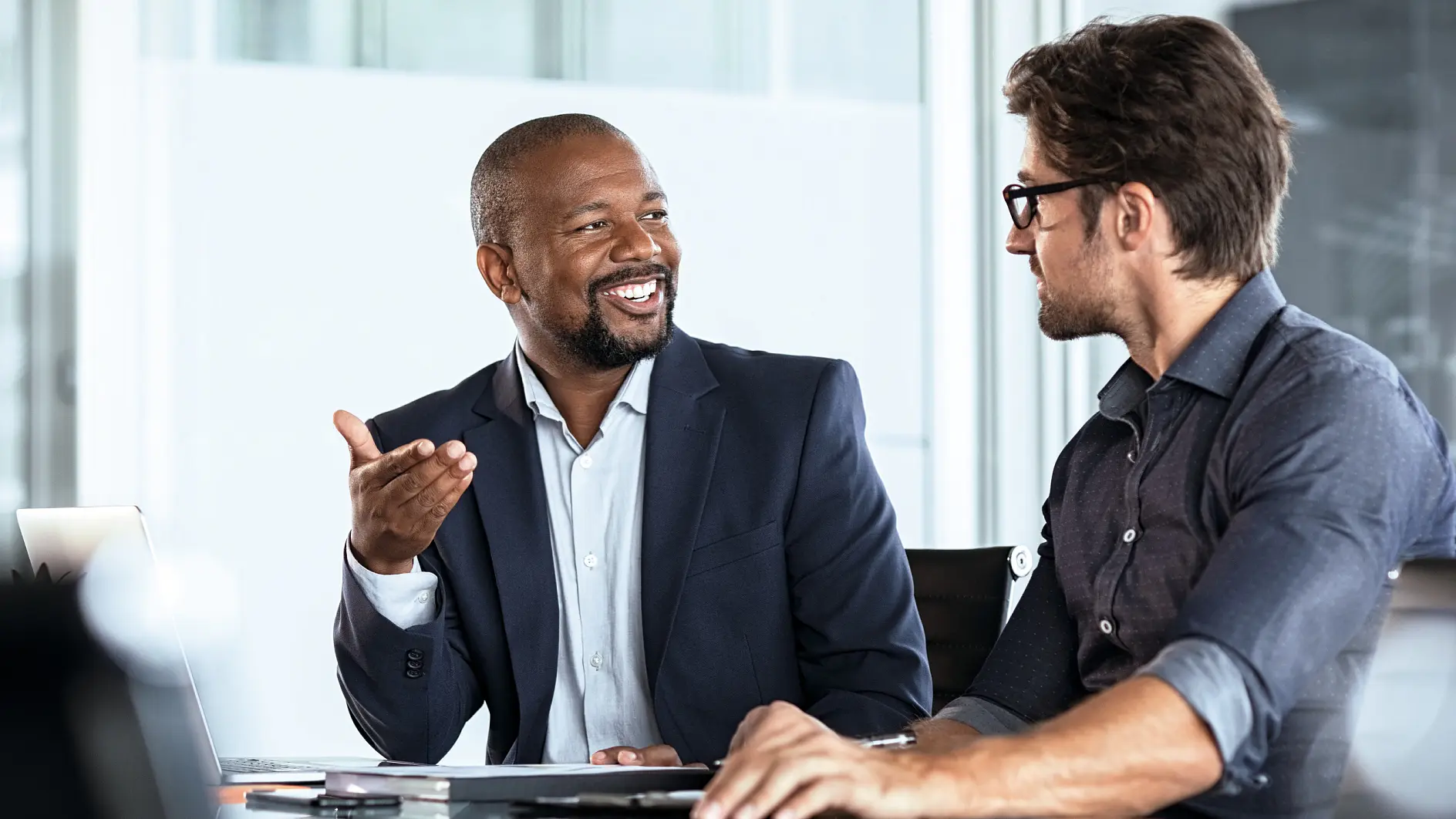 Two men in business attire engaged in discussion at an office desk with laptop and documents, indoor professional setting. (This text has been generated by AI)