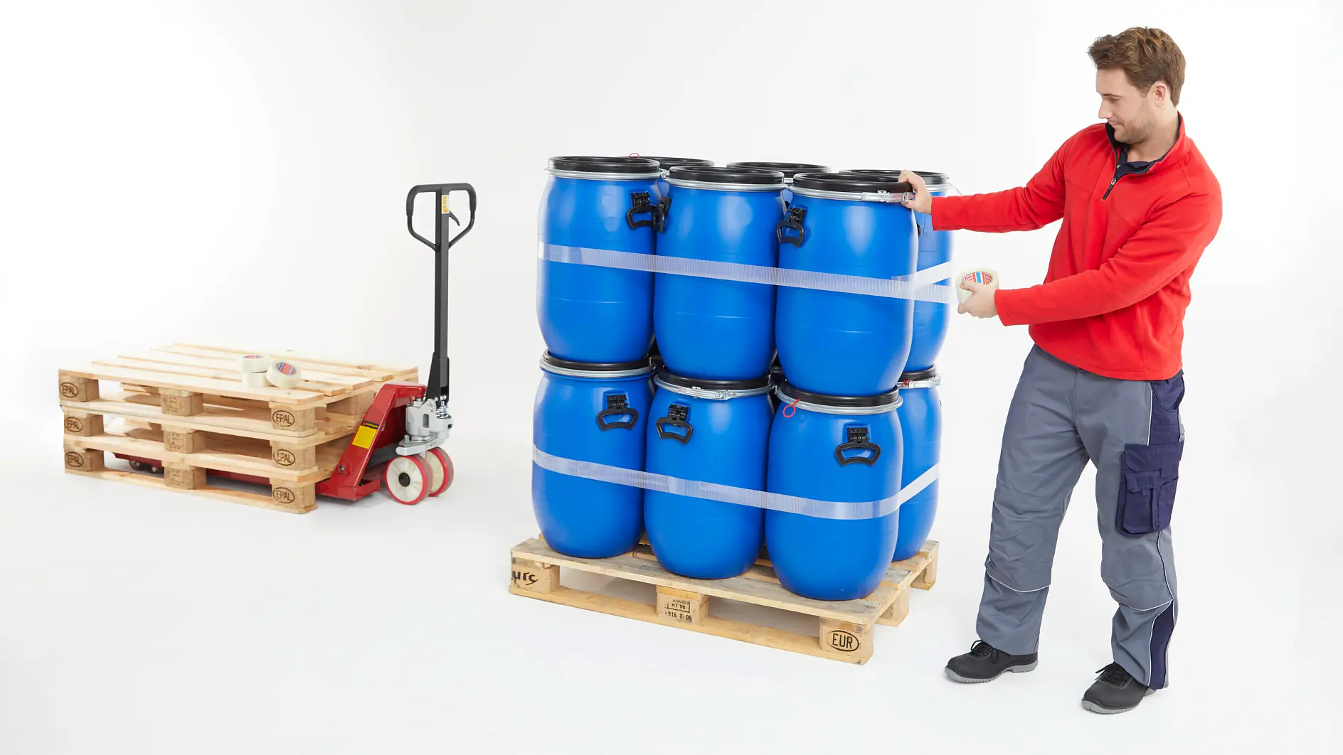 man in red jacket pushing wooden pallet stacked with six blue barrels indoors next to empty pallet and pallet jack tesa