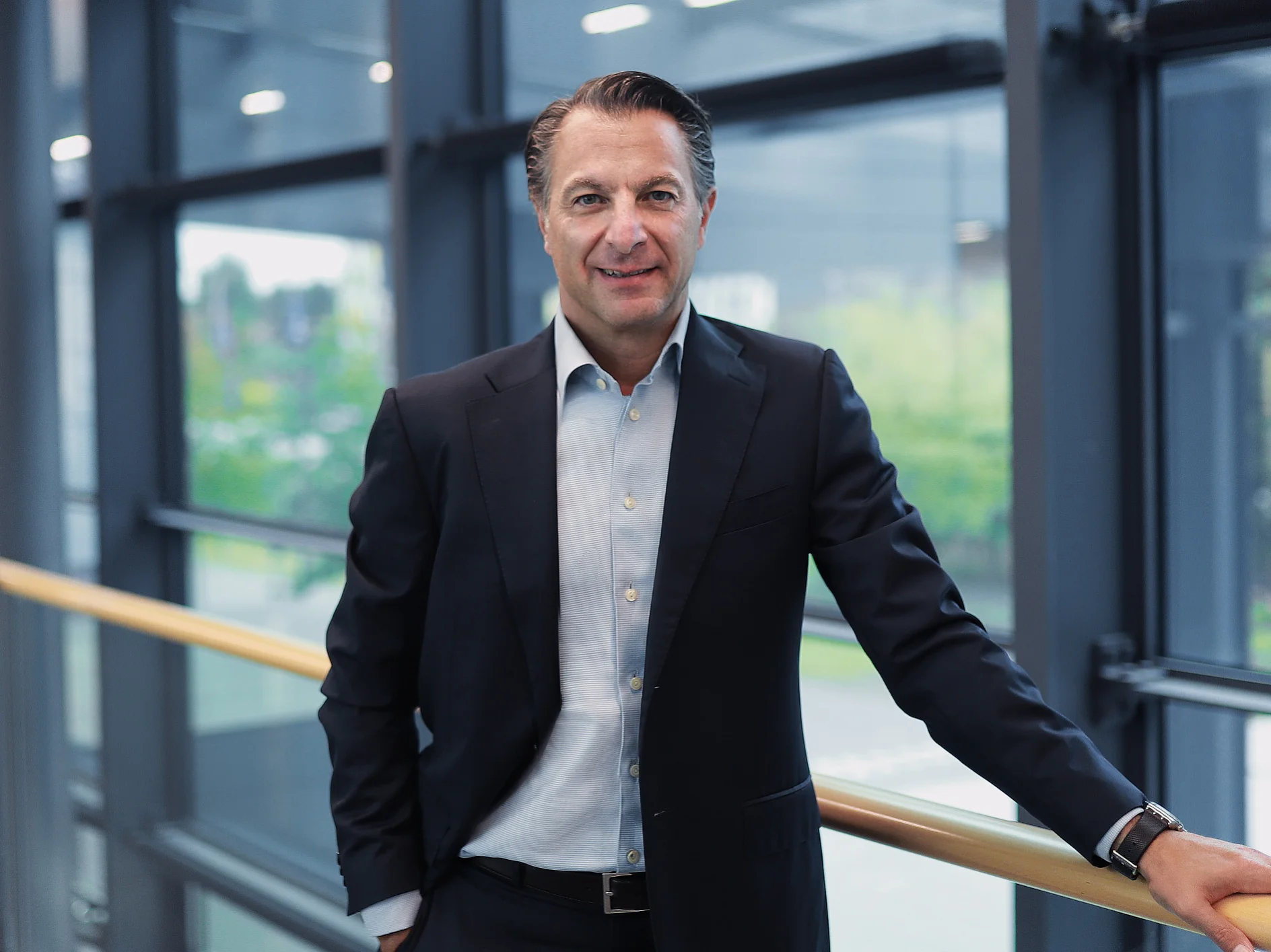 A man in a dark suit and light blue shirt stands by a railing indoors, with large windows and an outdoor view behind him, smiling slightly. (This text has been generated by AI)