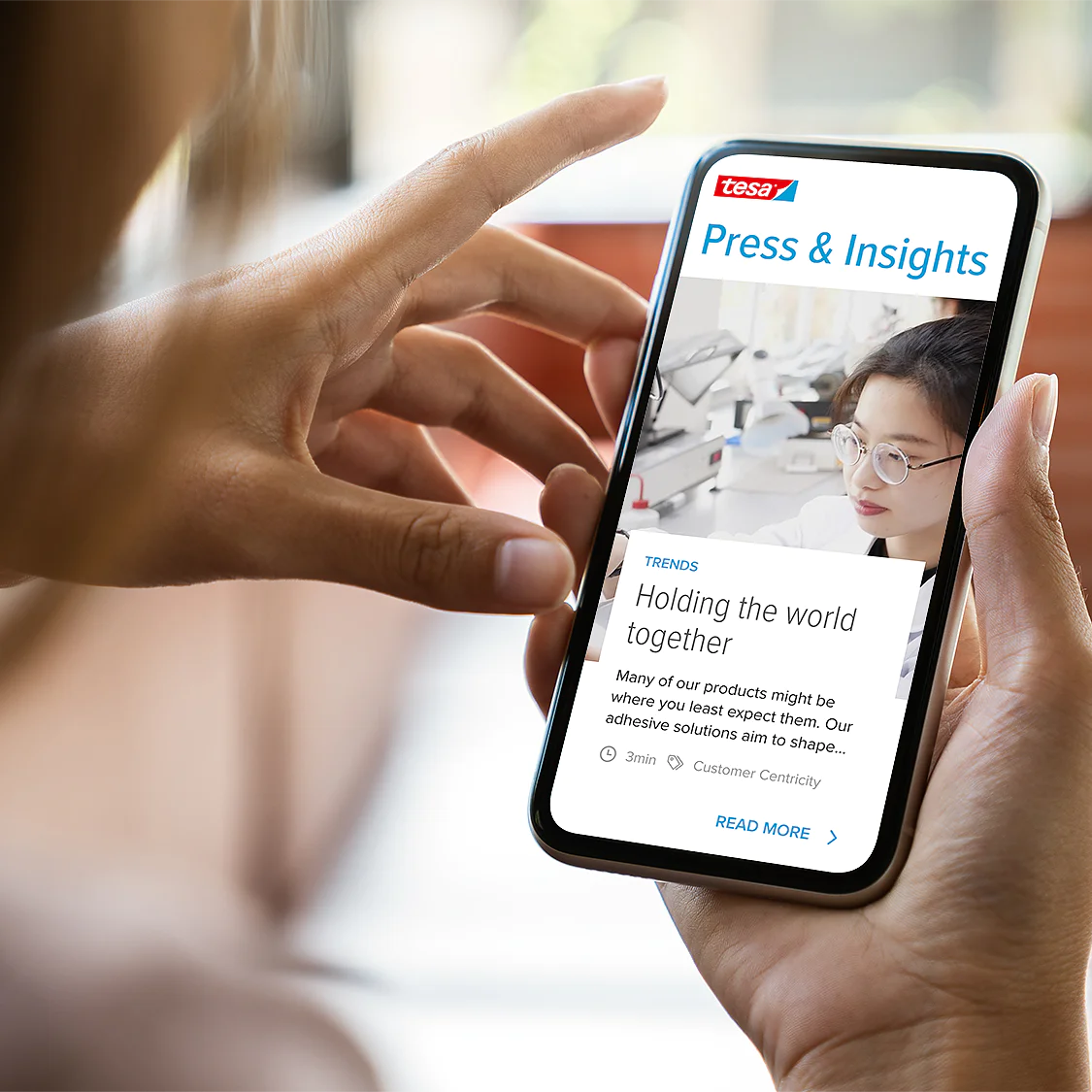 A person holding a smartphone displaying a webpage titled Press & Insights. The screen shows an article with an image of a woman behind a microscope and text about tesa tape adhesives. The smartphone is held in a hand with a blurred background. (This text has been generated by AI)