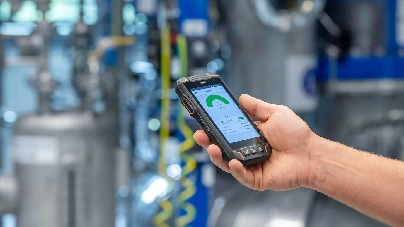 Uma pessoa segura um dispositivo portátil robusto com um medidor verde e dados exibidos, em uma instalação industrial com fita tesa nos tubos. (This text has been generated by AI)