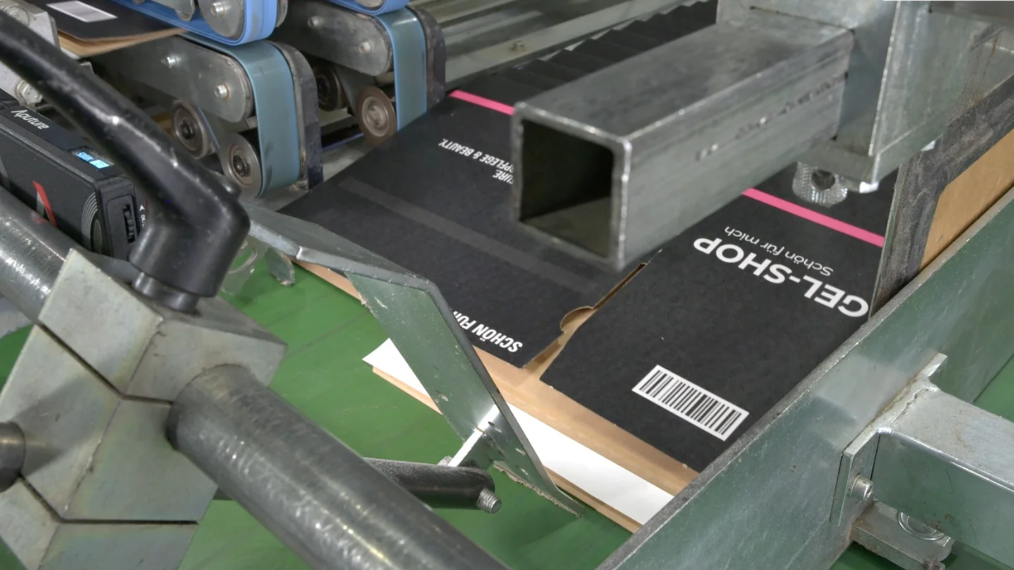 Um sistema de esteira com componentes metálicos está processando uma caixa de papelão rotulada como GEL-SHOP. A caixa está se movendo sob uma barra metálica, com roletes azuis visíveis ao fundo. O ambiente sugere um cenário de embalagem ou manufatura onde a fita tesa poderia ser utilizada de forma eficaz. (This text has been generated by AI)