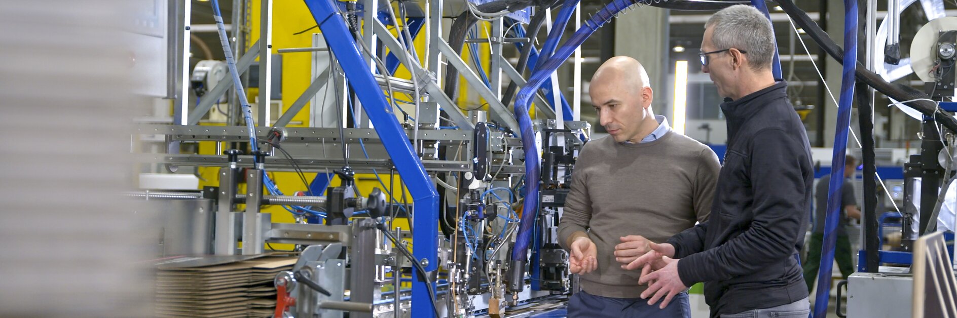 Dois homens estão em frente a máquinas industriais em uma fábrica. Eles parecem estar discutindo sobre o equipamento. Papelão ondulado é visível nas máquinas, fixado com fita tesa. O ambiente é bem iluminado e organizado, com vários componentes metálicos e azuis.
