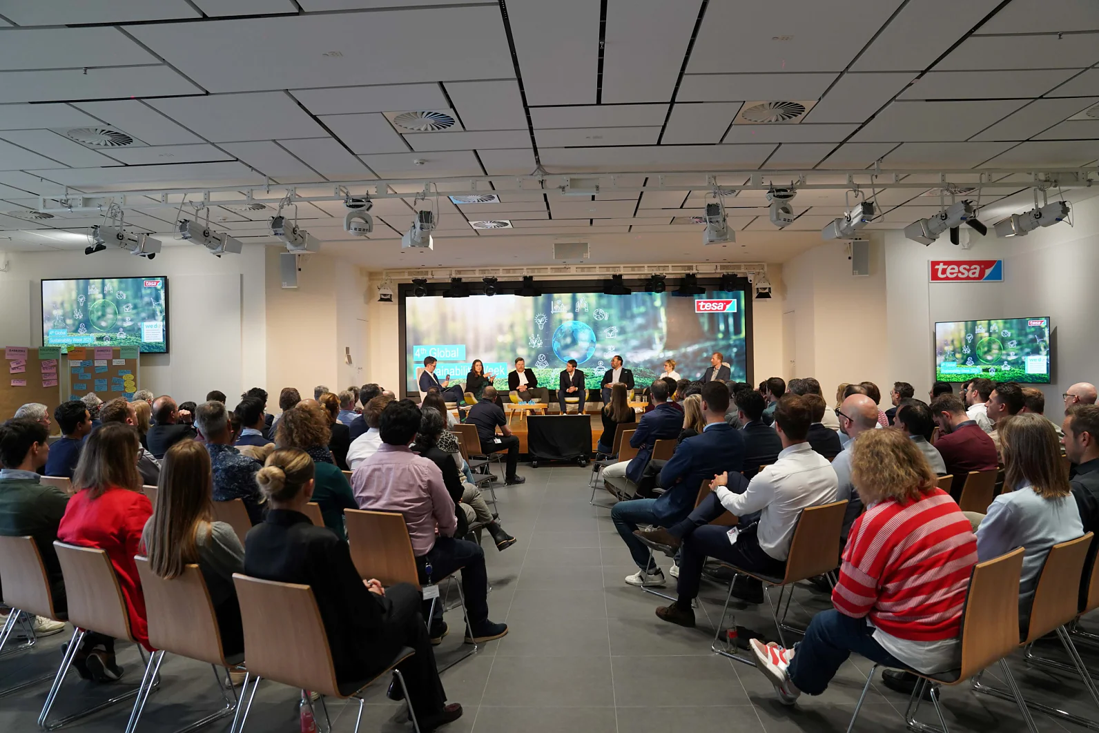 Uma plateia assiste a um painel em uma sala de conferências moderna. Os palestrantes estão sentados em uma mesa à frente; telas exibem o logo "tesa tape". (This text has been generated by AI)