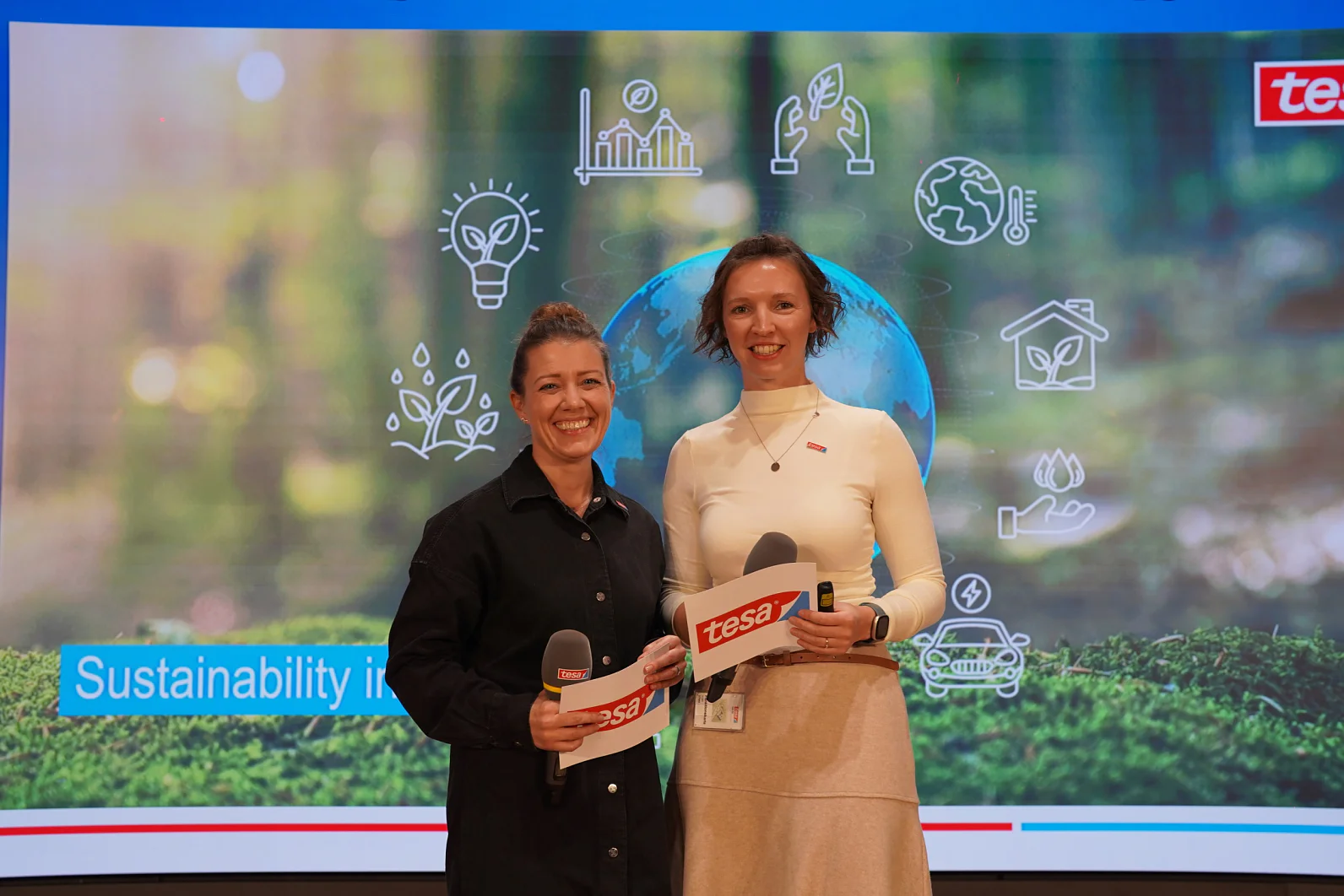 Duas mulheres estão lado a lado segurando microfones com o rótulo tesa tape em frente a uma tela com ícones de sustentabilidade e o texto Sustentabilidade em. (This text has been generated by AI)