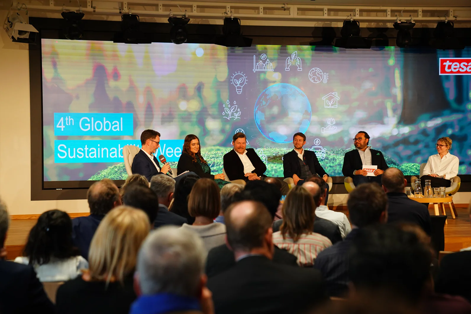 Painel de seis pessoas fala na 4ª Semana Global de Sustentabilidade; uma tela exibe gráficos do evento e o logo da tesa tape atrás deles. (This text has been generated by AI)