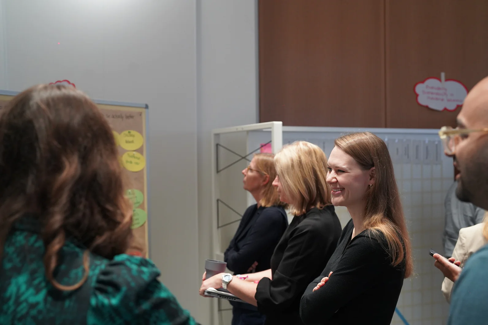 Um grupo de pessoas está em pé, dentro de um ambiente, algumas sorrindo e ouvindo, olhando para um quadro com notas circulares coloridas presas com fita tesa. (This text has been generated by AI)