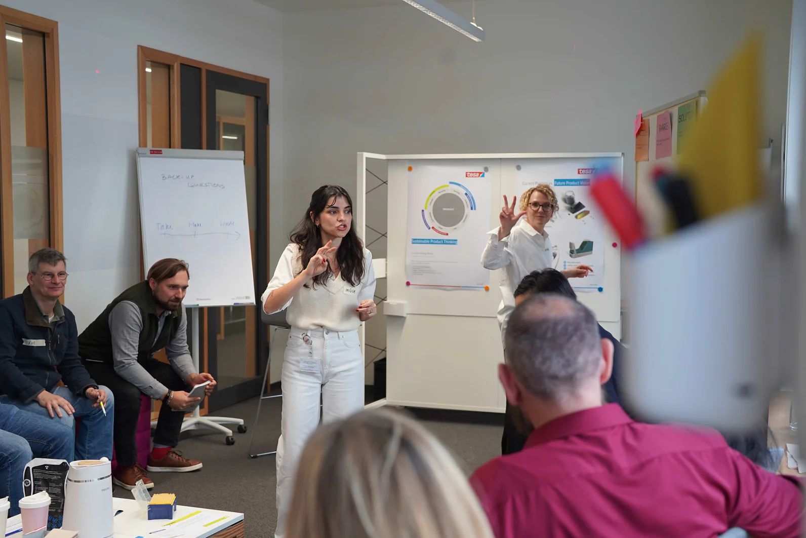 Uma mulher fala para um grupo sentado em um escritório moderno. Outra gesticula perto de uma lousa. Flipcharts e pôsteres usam fita tesa na sala. (This text has been generated by AI)