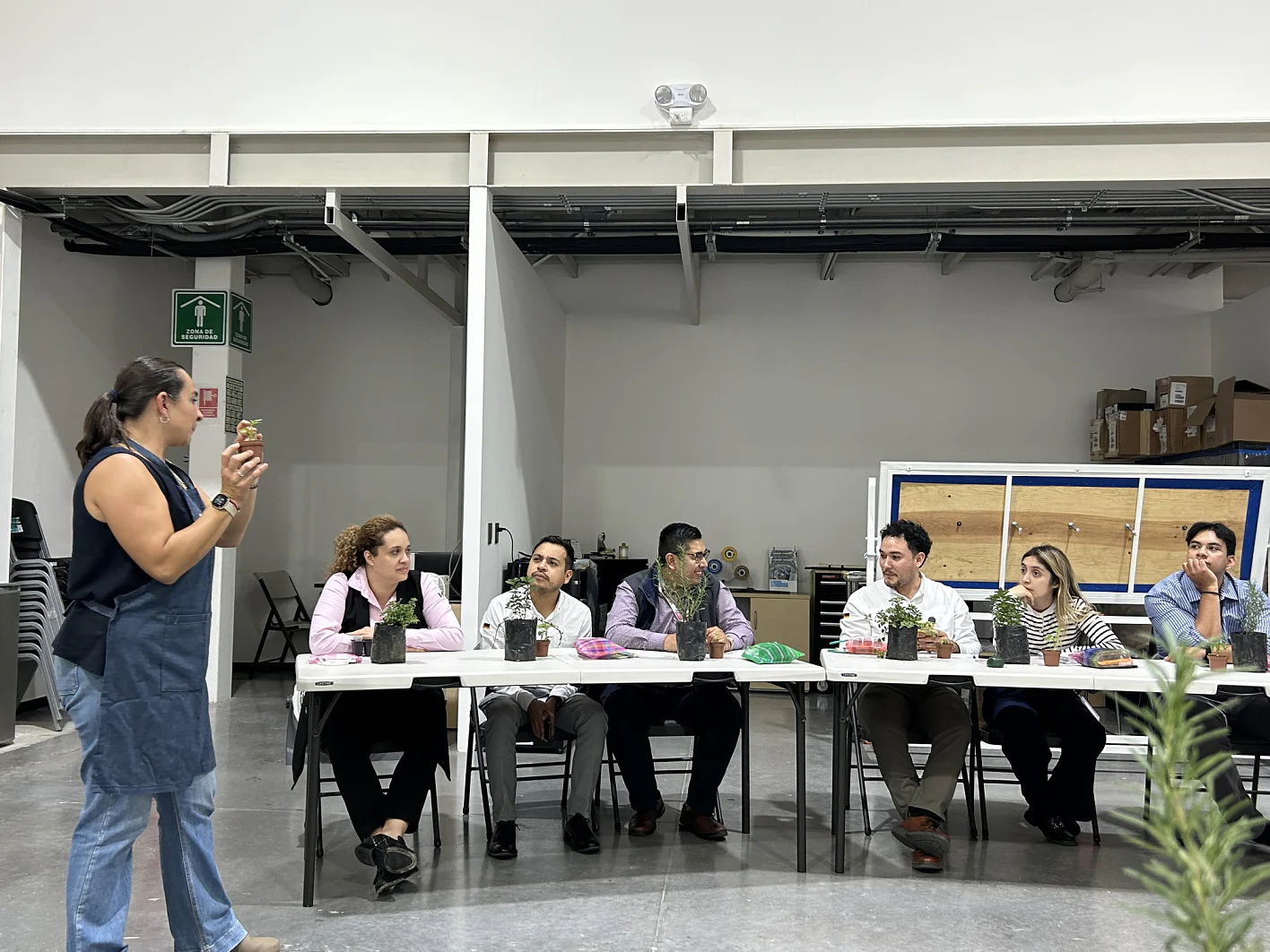 Uma mulher fala para seis pessoas em uma mesa comprida, cada uma com um vaso de planta. Prateleiras e cadeiras empilhadas estão ao fundo. (This text has been generated by AI)