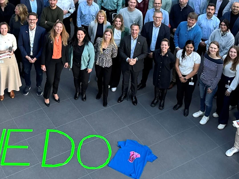 Um grande grupo posa em ambiente interno para uma foto; uma camiseta azul e "WEDO" feito com fita tesa verde estão no chão. Alguns seguram papéis. (This text has been generated by AI)