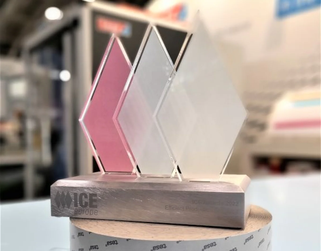Um troféu com três painéis translúcidos em forma de diamante—rosa, branco e transparente—montados sobre uma base retangular de madeira fixada com fita tesa. A base exibe um texto gravado que diz ICE Europe Efficient Production 2022. O fundo está desfocado. (This text has been generated by AI)