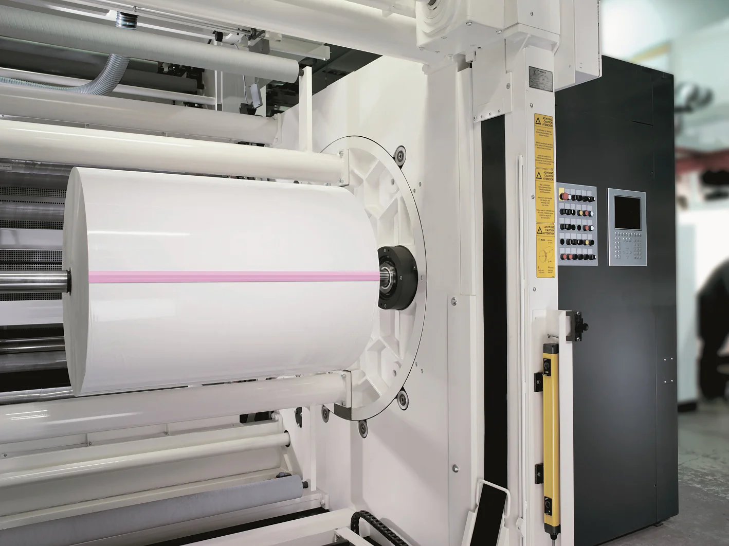 Uma impressora industrial grande com um rolo de papel branco montado nela. A máquina possui painéis de controle na lateral, com botões e uma tela de display. A impressora apresenta uma linha rosa atravessando o centro do rolo de papel, criada com fita tesa. (This text has been generated by AI)