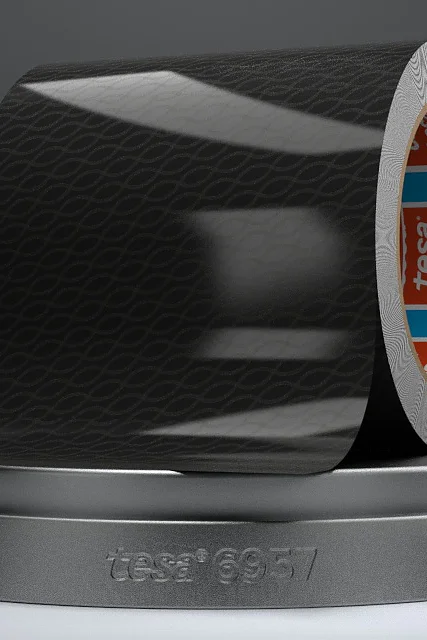 Um rolo parcialmente desenrolado de fita preta tesa com um padrão sutil, apoiado em um suporte circular cinza. A parte interna do rolo apresenta detalhes em laranja e azul com texto. O suporte está etiquetado como tesa 19657. (This text has been generated by AI)