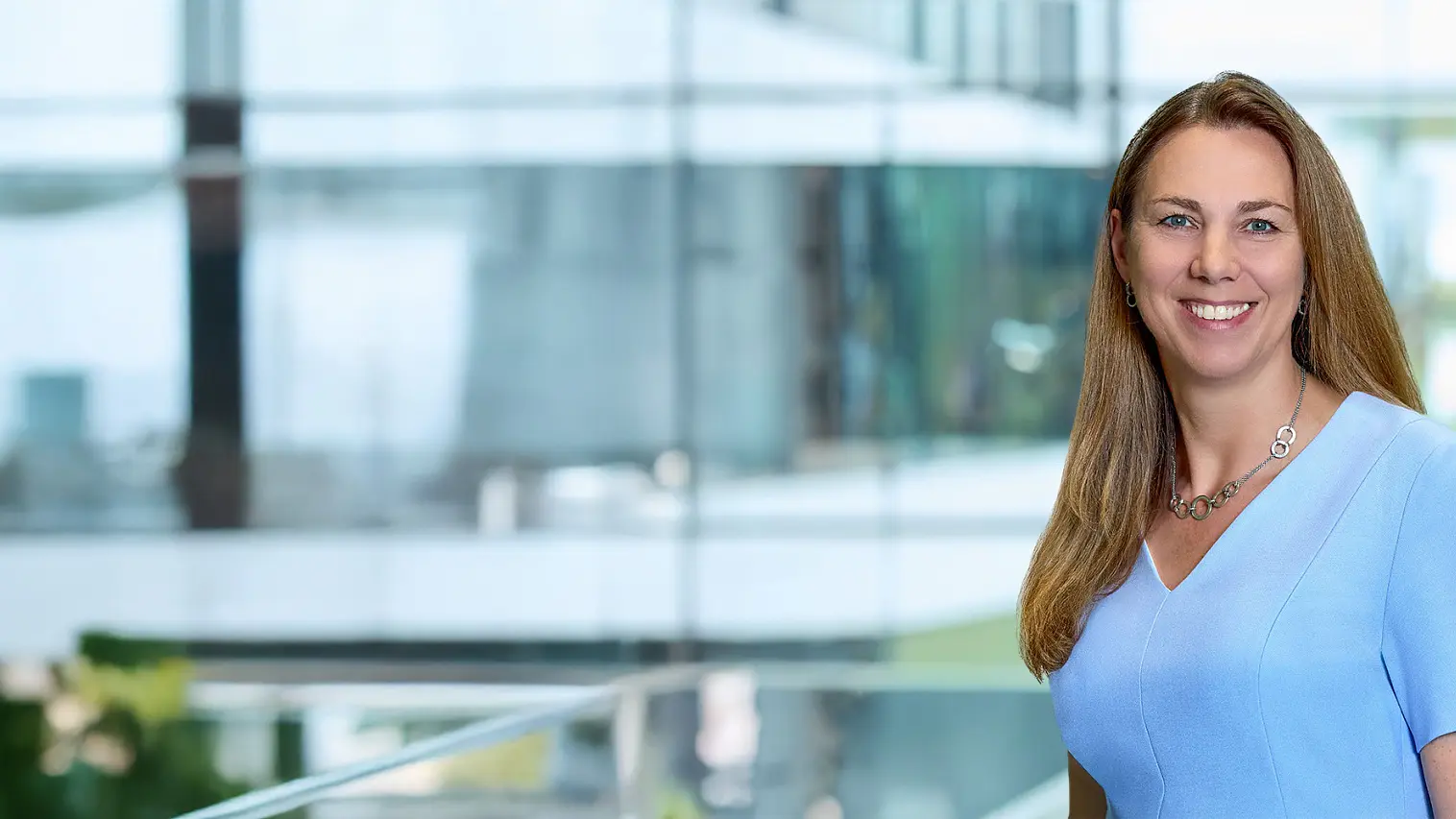 A woman with long brown hair, wearing a light blue short-sleeve top and a necklace, stands indoors by glass windows and green plants, smiling. (This text has been generated by AI)