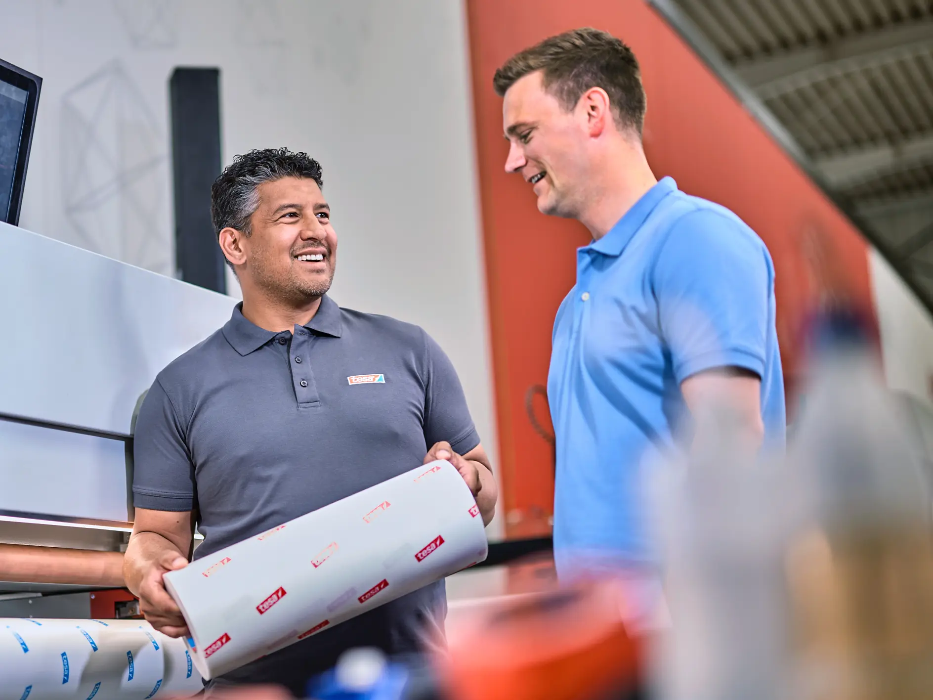 Dois homens estão ao lado de uma grande impressora em um ambiente interno. O homem de cinza segura um rolo de fita tesa, enquanto o homem de azul sorri para ele. (This text has been generated by AI)