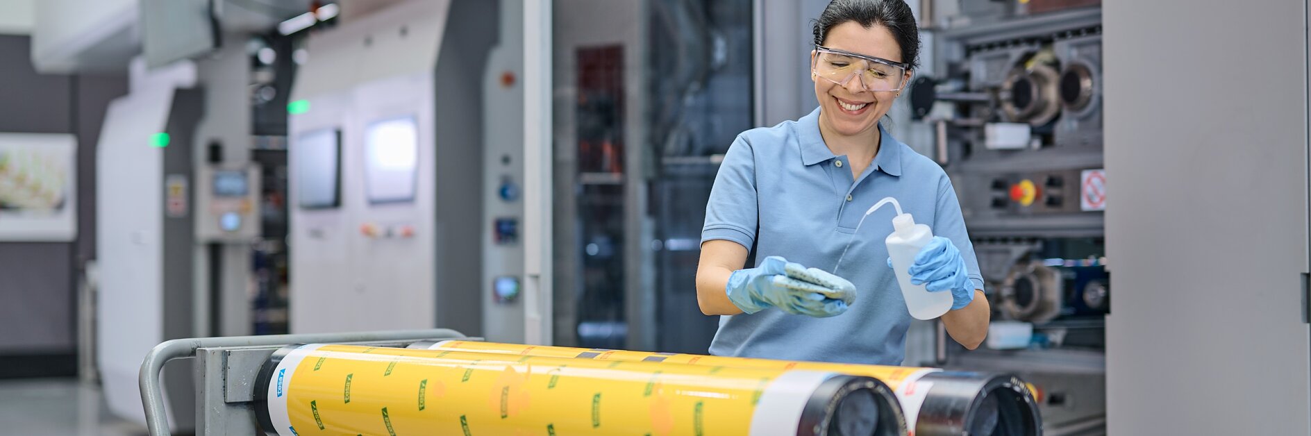 technician in blue uniform operating large industrial printing machine indoors, handling yellow printed material roll