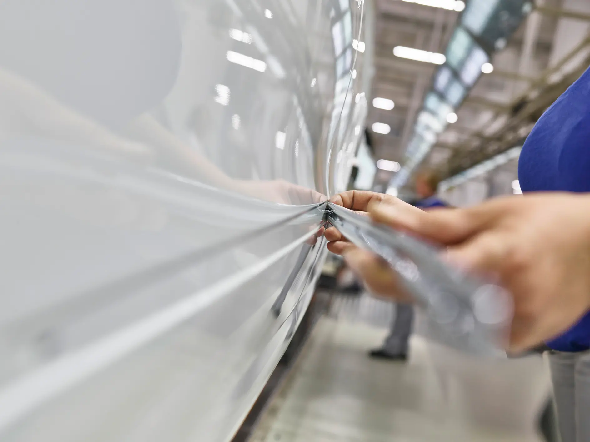 Fita tesa sendo aplicada em um veículo branco em ambiente fabril, com foco nas mãos alisando a fita sobre a superfície (This text has been generated by AI)