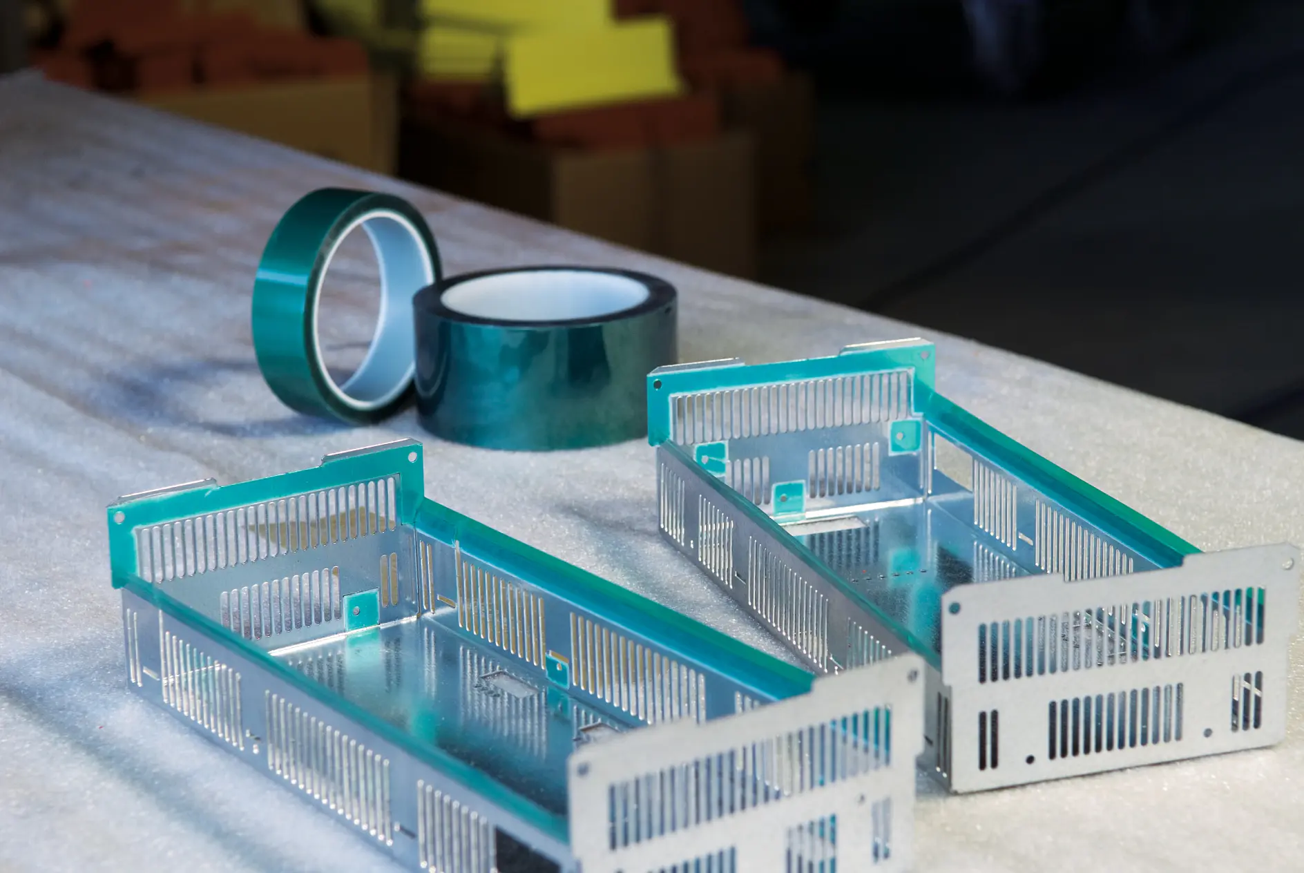 Dois quadros metálicos retangulares com ranhuras estão sobre uma superfície texturizada. Atrás deles há dois rolos de fita tesa verde. O fundo está desfocado, mostrando pilhas de papel ou papelão. (This text has been generated by AI)