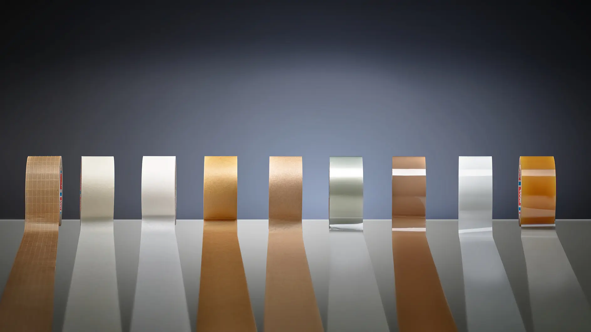 Rolos de fita adesiva tesa em diversas cores e materiais alinhados sobre uma superfície reflexiva contra um fundo escuro em degradê (This text has been generated by AI)