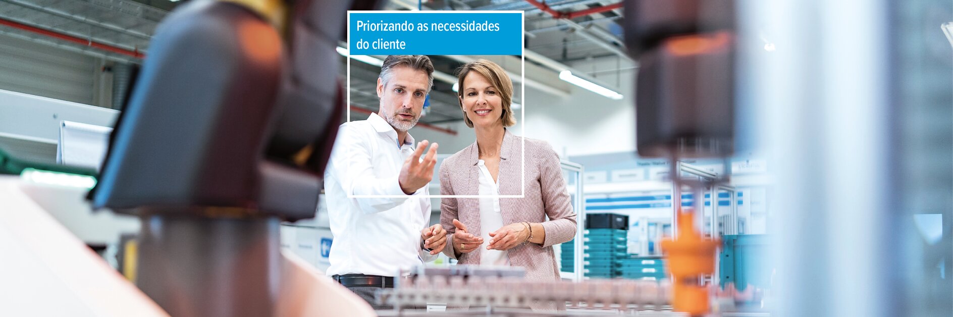 Duas pessoas em uma fábrica perto de um maquinário com uma sobreposição digital em português que diz: "Priorizando as necessidades do cliente".
