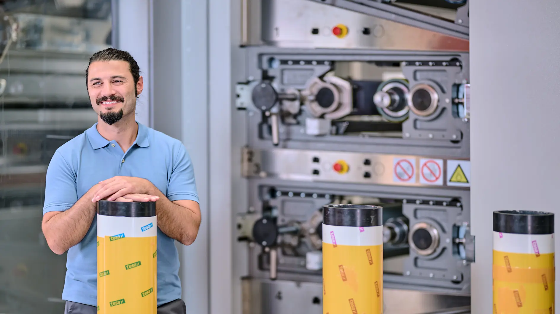Um homem com barba e cabelo preso em um rabo de cavalo, vestindo uma camisa polo azul, está sorrindo com as mãos sobre um grande objeto cilíndrico envolto em fita amarela da tesa. Máquinas industriais são visíveis ao fundo. (This text has been generated by AI)