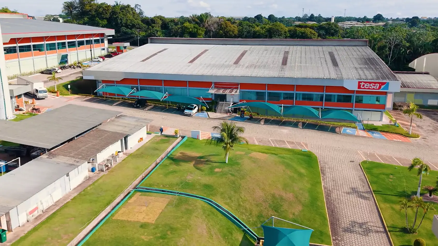 Um grande complexo de edifícios industriais com telhados vermelhos e cinzas, com a marca “tesa tape”, está cercado por áreas pavimentadas, veículos estacionados, árvores e gramados bem cuidados, fotografado de um ponto de vista elevado. (This text has been generated by AI)