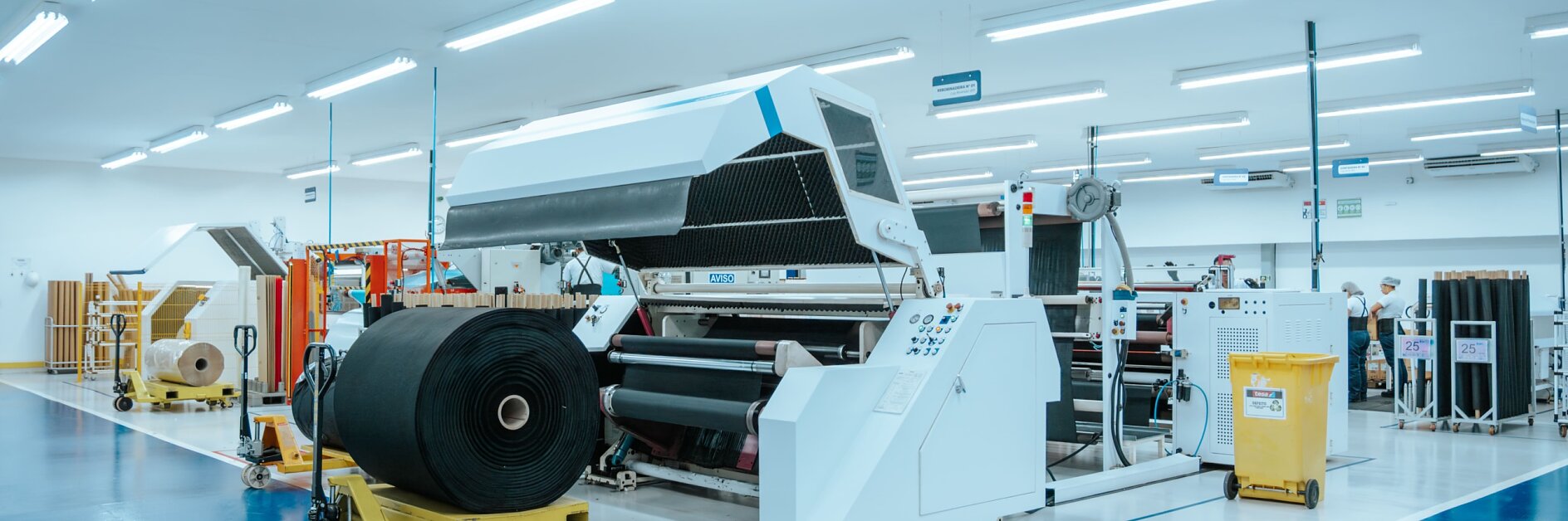 Uma grande máquina industrial processa um rolo de tecido preto em um espaço fabril limpo e bem iluminado, com piso azul e branco. Caixas amarelas e outros equipamentos, incluindo fita adesiva tesa, são visíveis ao fundo.