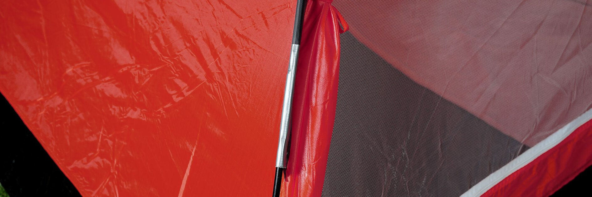 Close-up of a red and black camping tent, highlighting the seam where the fabric meets the zippered entrance. The tent fabric appears taut, with a mesh panel visible on the entrance. The ground beneath is covered in green grass. (This text has been generated by AI)