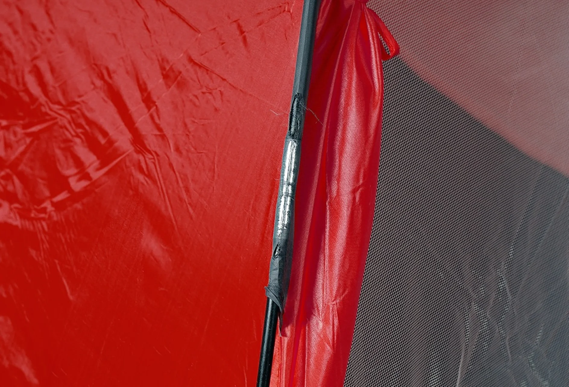 Close-up of a red tents fabric with visible wear at the seam line, showing black pole support. The fabric appears slightly frayed, revealing a silver section on the pole. The background is partially visible mesh material secured with tesa tape for added reinforcement. (This text has been generated by AI)