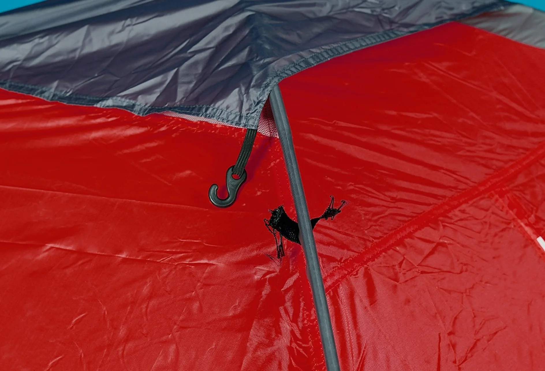 A close-up of a red tent with a tear in the fabric. A black hook is visible attached to a tent pole, which supports the structure. The tear is located near the hook and is about halfway up the tent wall. The fabric appears slightly wrinkled, but using tesa tape can effectively mend it and provide added durability. (This text has been generated by AI)