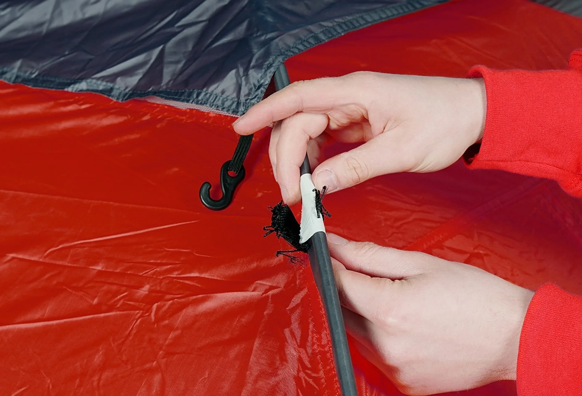 Hands are assembling a tent by attaching a black hook and loop strap to a pole over red and gray tent fabric using tesa tape. The person is wearing a red garment. (This text has been generated by AI)