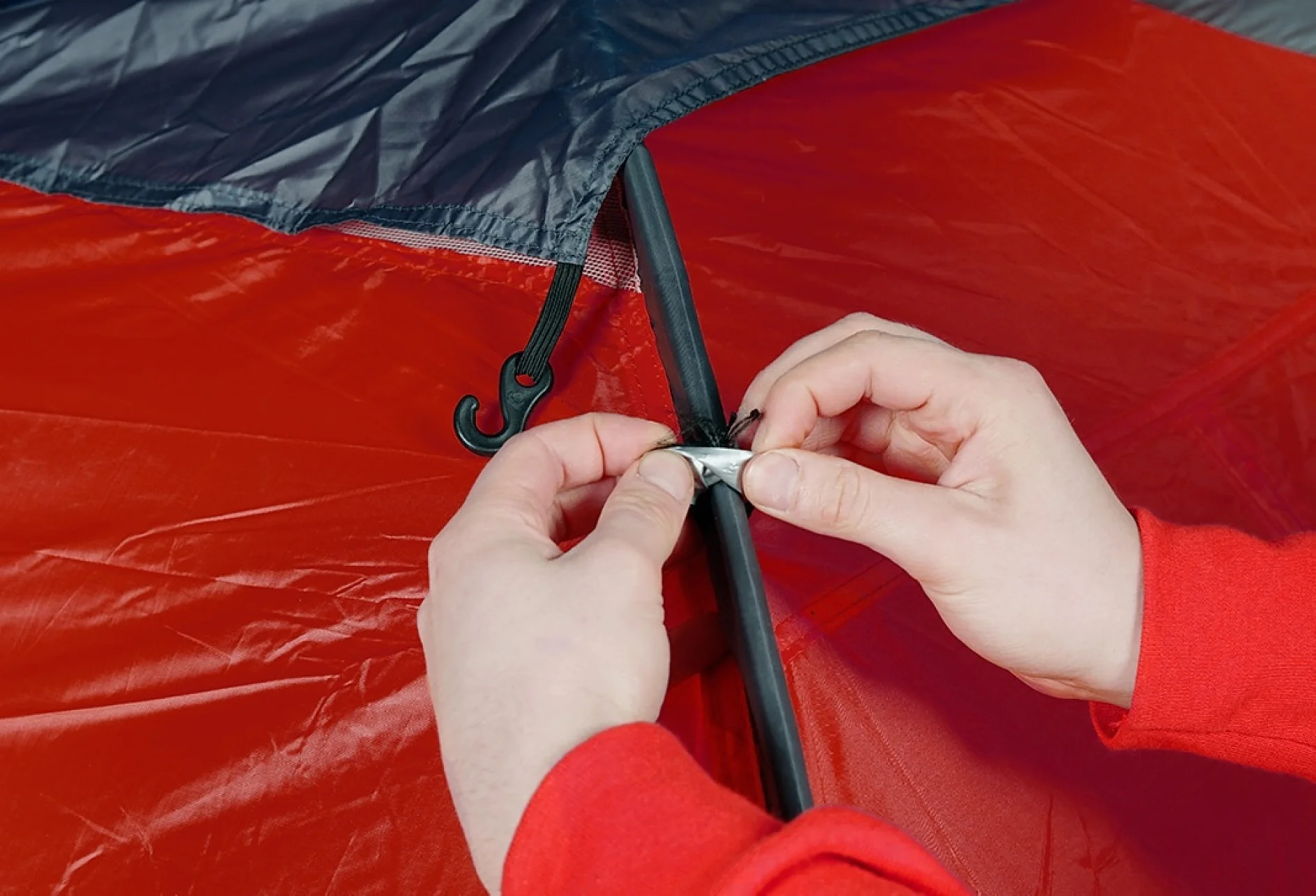 Two hands are assembling a tent, attaching a black pole to a red fabric with a tesa tape fastener. The tent has a black and red color scheme. The person is wearing a red long-sleeve shirt. (This text has been generated by AI)