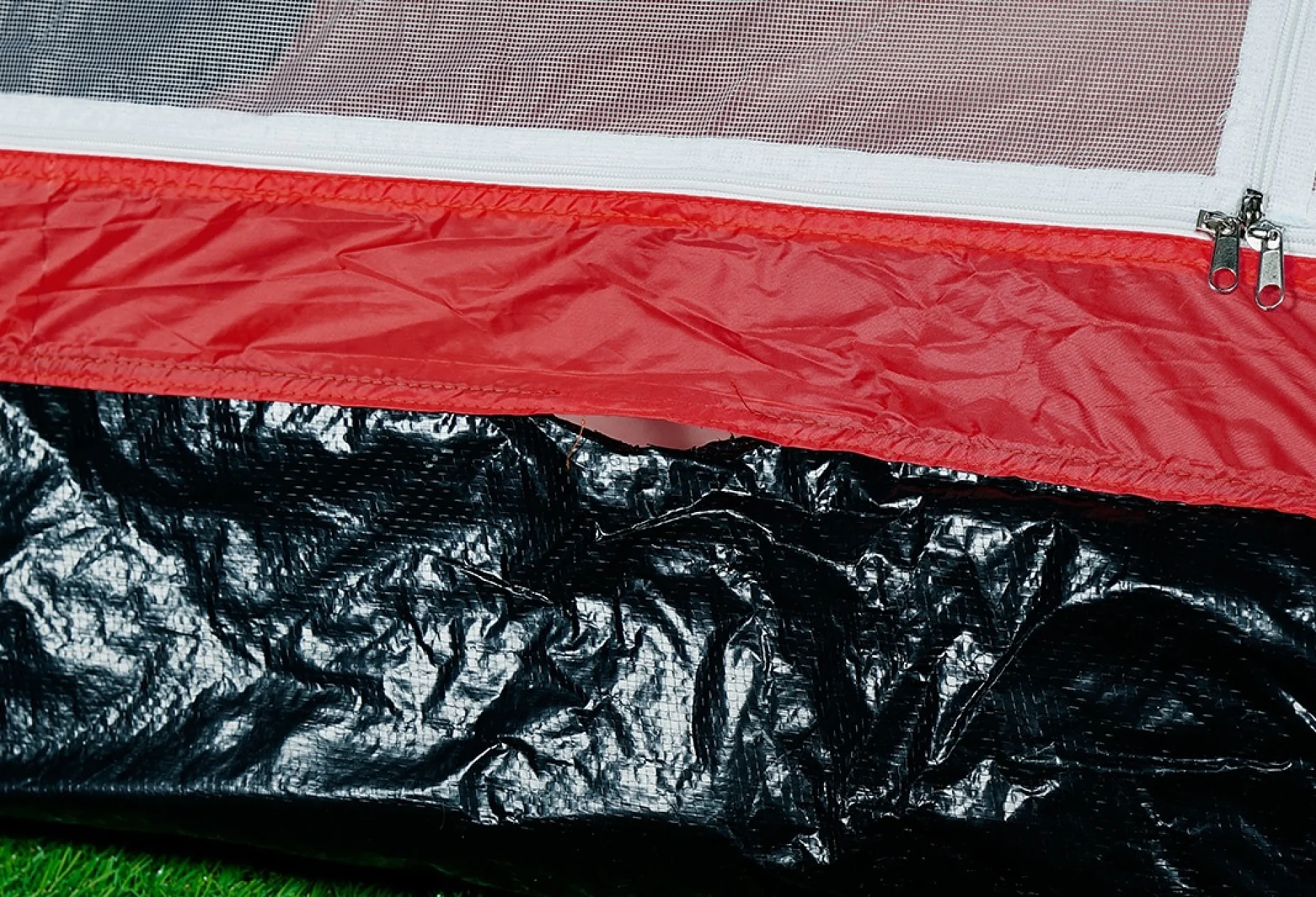 Close-up of a camping tent’s edge showing the red fabric with a zipper and white mesh above. Below, the base is made of crinkled black material, set on a patch of green grass. (This text has been generated by AI)